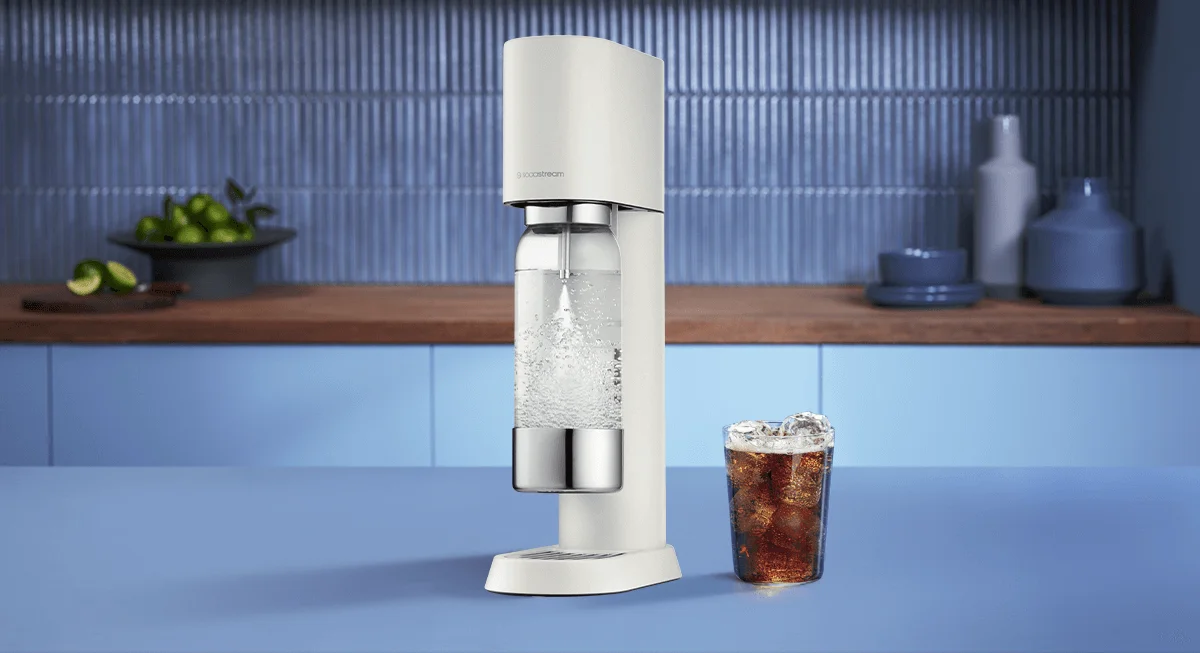 Saturator Sodastream Enso oraz szklanka z napojem gazowanym na tle niebieskiej kuchni